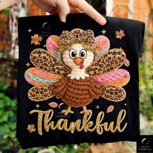 May include: Black t-shirt featuring a crocheted turkey design with a leopard print bow and wings, and the word "Thankful" in gold glitter lettering. The turkey has a white head, brown body, and colorful wings. Autumn-themed leaves are scattered around the design.