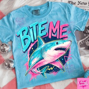 Puede incluir: Camiseta azul claro con un gr&aacute;fico de un tibur&oacute;n y las palabras "BITE ME" en turquesa y rosa. El tibur&oacute;n es en tonos de azul y rosa, con una boca blanca y dientes afilados. La camiseta es de material suave.