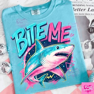 Puede incluir: Camiseta azul claro con un gr&aacute;fico de tibur&oacute;n y las palabras "BITE ME" en turquesa y rosa. El tibur&oacute;n est&aacute; detallado con tonos de azul y rosa, sobre un fondo azul oscuro y amarillo. La camiseta tiene cuello redondo.