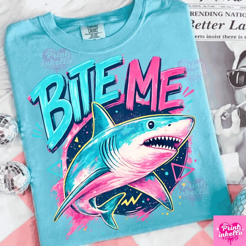 Puede incluir: Camiseta azul claro con un gr&aacute;fico de tibur&oacute;n y las palabras "BITE ME" en turquesa y rosa. El tibur&oacute;n est&aacute; detallado con tonos de azul y rosa, sobre un fondo azul oscuro y amarillo. La camiseta tiene cuello redondo.