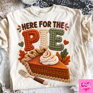 Op de afbeelding: Een crèmekleurig t-shirt met een afbeelding van een stuk pompoentaart met slagroom, een houten vork en herfstbladeren. De tekst HERE FOR THE PIE is in bruin, oranje en groen, met een textuur die eruitziet als gebreid garen.