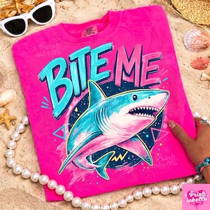Puede incluir: Una camiseta rosa intenso con las palabras "BITE ME" en turquesa y rosa. Un tibur&oacute;n de dibujos animados con colores azul y rosa est&aacute; centrado debajo del texto. La camiseta es de cuello redondo.