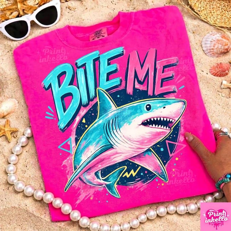 Puede incluir: Una camiseta rosa intenso con las palabras "BITE ME" en turquesa y rosa. Un tibur&oacute;n de dibujos animados con colores azul y rosa est&aacute; centrado debajo del texto. La camiseta es de cuello redondo.