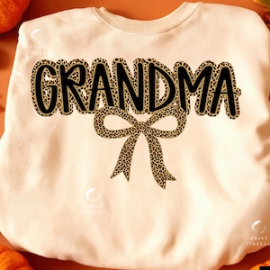 May include: Cream-colored sweatshirt with the word "GRANDMA" in large, black, leopard print letters. A leopard print bow is centered below the word. The sweatshirt is a casual, comfortable style.