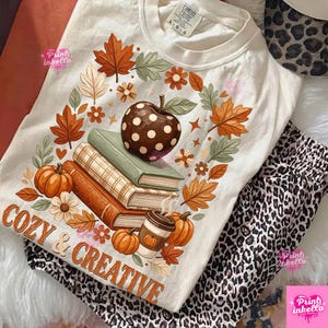 May include: A cream-colored t-shirt featuring a graphic of a polka-dot apple resting on a stack of two books, a coffee cup, and pumpkins, all surrounded by autumn leaves and flowers. The text COZY & CREATIVE is printed in orange at the bottom.