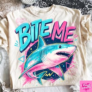 Puede incluir: Camiseta color crema con un gr&aacute;fico de tibur&oacute;n colorido. El dise&ntilde;o incluye las palabras "BITE ME" en turquesa y rosa, con una ilustraci&oacute;n detallada de un tibur&oacute;n en tonos azules y rosas.