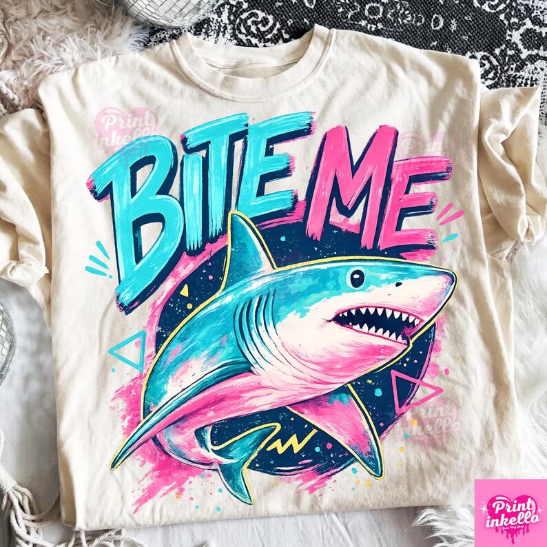 Puede incluir: Camiseta color crema con un gr&aacute;fico de tibur&oacute;n colorido. El dise&ntilde;o incluye las palabras "BITE ME" en turquesa y rosa, con una ilustraci&oacute;n detallada de un tibur&oacute;n en tonos azules y rosas.