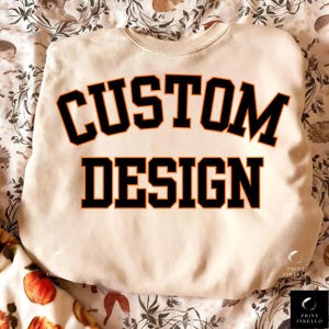 May include: Cream-colored sweatshirt with the words "CUSTOM DESIGN" in large, black, block letters with orange outlines. The sweatshirt is displayed on a patterned fabric background.