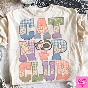 Könnte beinhalten: Ein cremefarbenes T-Shirt mit dem Schriftzug CAT NAP CLUB in großen Blockbuchstaben. Die Buchstaben sind mit verschiedenen pastellfarbenen Mustern gefüllt, darunter Vichy-Karo, Blumen und Punkte. Eine kleine Illustration einer schlafenden Katze und ein Pfotenabdruck sind zwischen den Buchstaben zentriert.