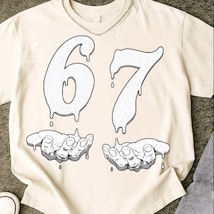 May include: Cream-colored t-shirt featuring a graphic design. The design includes the numbers "67" in a dripping style, with each number above a pair of hands. The hands are cupped, with drips falling from the numbers into the hands. A silver chain is around the neck.