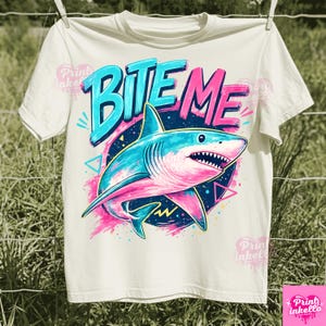 Puede incluir: Camiseta blanca con un gr&aacute;fico de un tibur&oacute;n y las palabras "BITE ME". El tibur&oacute;n es azul y rosa, sobre un c&iacute;rculo azul oscuro con detalles en rosa. El texto "BITE ME" es en azul y rosa. La camiseta est&aacute; colgada en un tendedero.