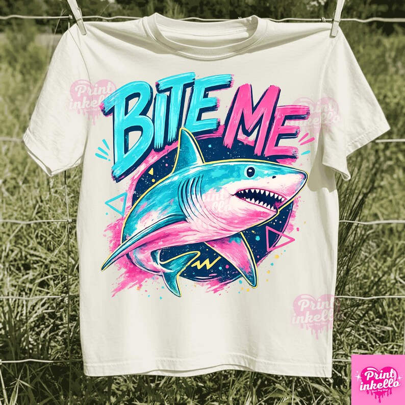 Puede incluir: Camiseta blanca con un gr&aacute;fico de un tibur&oacute;n y las palabras "BITE ME". El tibur&oacute;n es azul y rosa, sobre un c&iacute;rculo azul oscuro con detalles en rosa. El texto "BITE ME" es en azul y rosa. La camiseta est&aacute; colgada en un tendedero.