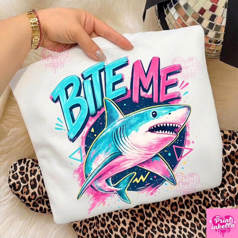 Puede incluir: Sudadera blanca con un gr&aacute;fico colorido de un tibur&oacute;n y las palabras "BITE ME" en azul y rosa. El tibur&oacute;n es azul y rosa, con vientre blanco y dientes afilados. El dise&ntilde;o incluye formas geom&eacute;tricas y un fondo azul oscuro.