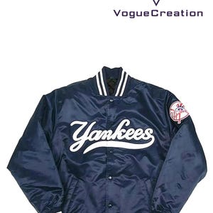 Può includere: Una giacca bomber in satin blu navy con bordini a costine bianchi e un logo "Yankees" bianco sul davanti. La giacca ha una chiusura con bottoni e una piccola toppa con il logo dei New York Yankees sulla manica destra.