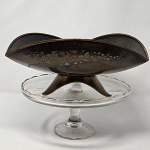 May include: A vintage brass bowl with an intricate cut-out design sits on a glass cake stand with a gold rim. The bowl has a dark brown patina and the stand has a clear glass base.