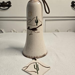 Puede incluir: Una campana de cerámica blanca con un asa de cuerda marrón. La campana está decorada con una escena del desierto con un correcaminos, un cactus y un sol. Una etiqueta de cerámica blanca más pequeña con un diseño de cactus está unida a la campana.
