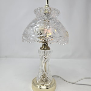 May include: A clear crystal table lamp with a floral pattern on the shade and base. The lamp has a gold-colored base and accents, and a white power cord. The lamp is approximately 12 inches tall.