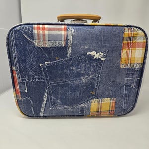 Peut inclure: Une valise vintage avec un motif patchwork en denim. La valise présente une poignée en bois et un mélange de denim bleu et de patchs de tissu à carreaux rouges, jaunes et blancs. La valise est de forme rectangulaire avec des coins arrondis.