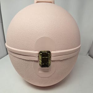 May include: A light pink, spherical vintage travel case with a textured surface. The case has a handle on top, a hinged lid, and a brass-colored clasp. The case is closed and appears to be empty.