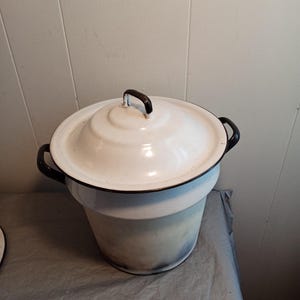 May include: A white enamel pot with a matching lid. The pot has black handles and a black handle on the lid. The pot is sitting on a gray surface. The pot is a vintage kitchenware item.