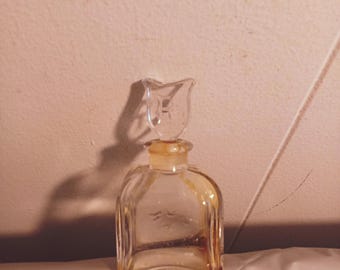 May include: A clear glass bottle with a textured surface and a clear glass stopper. The bottle has a square shape and is filled with a light yellow liquid.