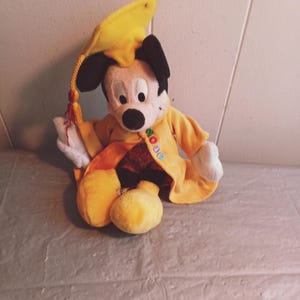 Puede incluir: Un peluche de Mickey Mouse con birrete y toga amarillos. El birrete tiene una borla negra y la toga tiene bordada la palabra "NO". El juguete est&aacute; sentado sobre una superficie gris.
