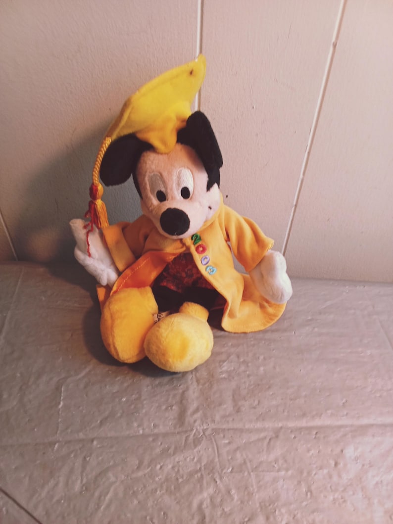 Puede incluir: Un peluche de Mickey Mouse con birrete y toga amarillos. El birrete tiene una borla negra y la toga tiene bordada la palabra "NO". El juguete est&aacute; sentado sobre una superficie gris.