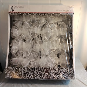 Puede incluir: Un paquete de 12 lazos de boda de tul blanco. Los lazos están empaquetados en una envoltura de plástico transparente con una caja de patrón blanco y negro. La caja tiene el texto "Wedding Bows 12 Count".