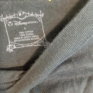 May include: Close-up of a dark gray t-shirt with a ribbed collar. The Disney Store logo is visible, along with text indicating the shirt is 100% cotton and made in South Africa. A yellow tag is attached to the shirt.