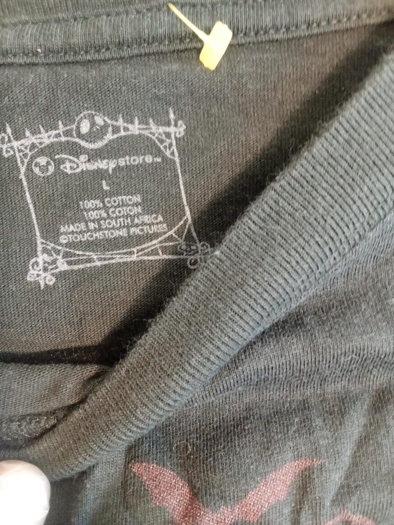 May include: Close-up of a dark gray t-shirt with a ribbed collar. The Disney Store logo is visible, along with text indicating the shirt is 100% cotton and made in South Africa. A yellow tag is attached to the shirt.