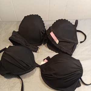May include: Four black bikini tops are displayed. Two tops have a scalloped edge, and the other two have a gathered fabric design. One top has a gold-colored clasp. All tops have straps and are made of a smooth fabric.
