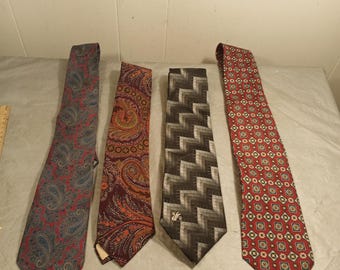 May include: Four neckties with various patterns are displayed. The ties include a paisley design in red and blue, a paisley design in brown and orange, a geometric zigzag pattern in black and gray, and a red tie with a repeating floral pattern.