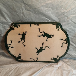 Puede incluir: Una bandeja de cerámica rectangular con bordes redondeados y festoneados que presenta una superficie de color crema con motas de color marrón oscuro. La bandeja está decorada con múltiples siluetas de ranas verde oscuro.