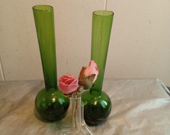 May include: Two green glass vases with a bulbous base and tall, slender necks. A small, clear glass vase holds two pink roses. The vases are arranged on a neutral-toned surface.