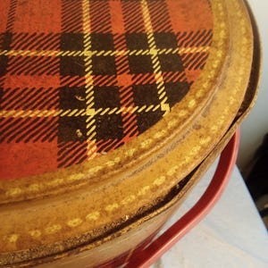 May include: Vintage metal picnic basket with a red, black, and yellow plaid pattern on the lid. The basket has a tan-colored body and a red plastic handle. The lid is slightly ajar, revealing the interior.