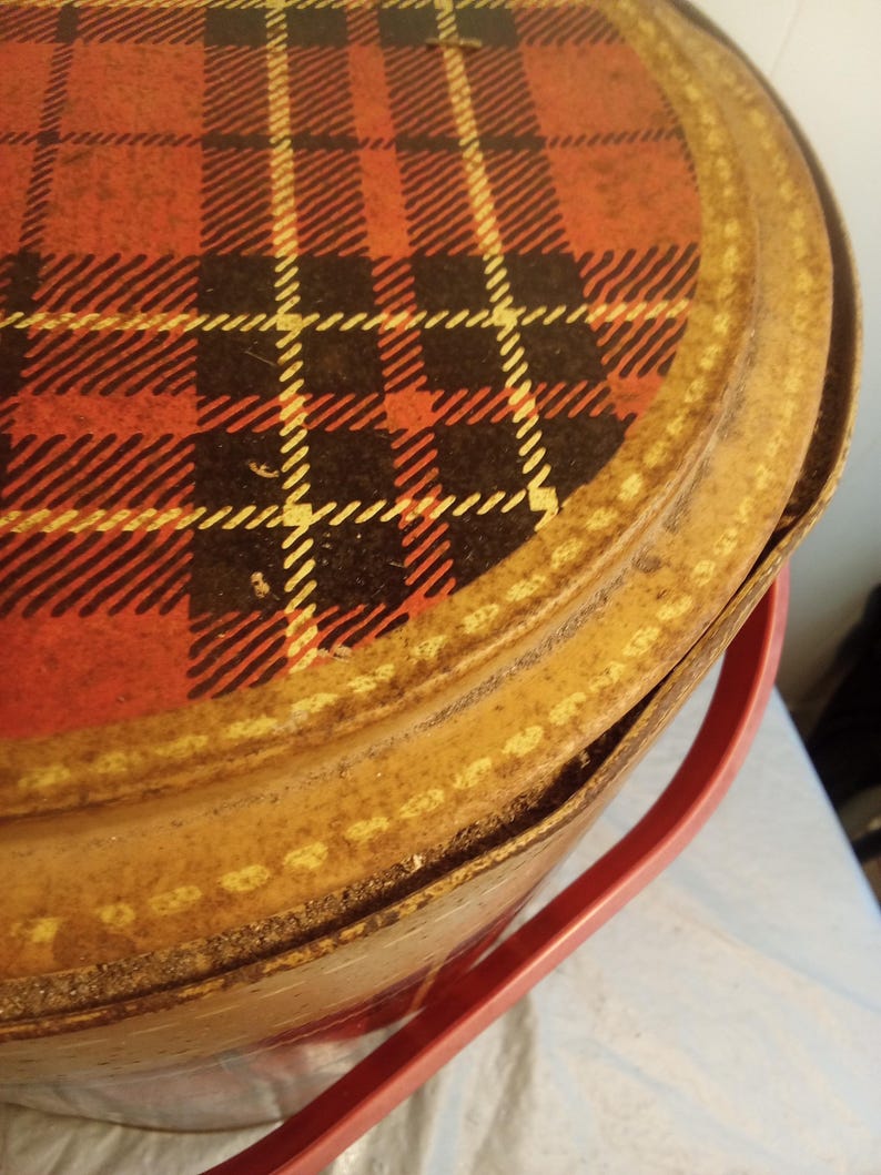 May include: Vintage metal picnic basket with a red, black, and yellow plaid pattern on the lid. The basket has a tan-colored body and a red plastic handle. The lid is slightly ajar, revealing the interior.