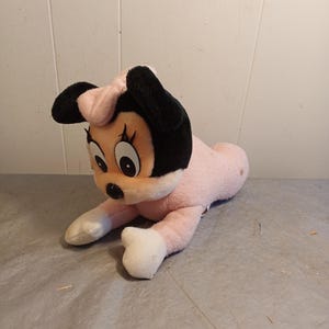 May include: A plush Minnie Mouse toy in a lying down pose. The toy features a pink body, white paws, and a black head with large eyes and ears. A pink bow sits atop the head. The toy is made of soft, fuzzy material.