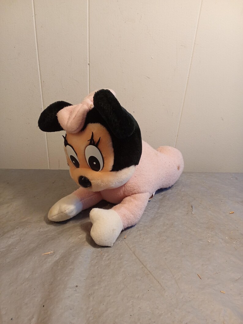 May include: A plush Minnie Mouse toy in a lying down pose. The toy features a pink body, white paws, and a black head with large eyes and ears. A pink bow sits atop the head. The toy is made of soft, fuzzy material.