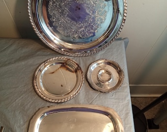 May include: A collection of silver serving trays and dishes. The largest tray is round with a decorative edge and ornate center design. Smaller round and oval trays are also present, all with a shiny, reflective surface. The items are arranged on a gray surface.
