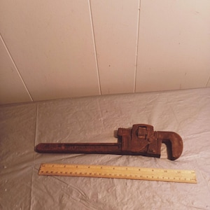 May include: A rusty brown pipe wrench with a long handle and adjustable jaws. A wooden ruler is placed next to the wrench for scale. The tool is designed for gripping and turning pipes.