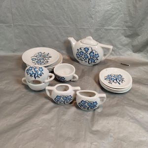 Peut inclure: Un service à thé en céramique blanche avec des motifs floraux bleus. L'ensemble comprend une théière, des tasses, des soucoupes et de petits récipients à crème et à sucre. Le service est posé sur une surface grise.