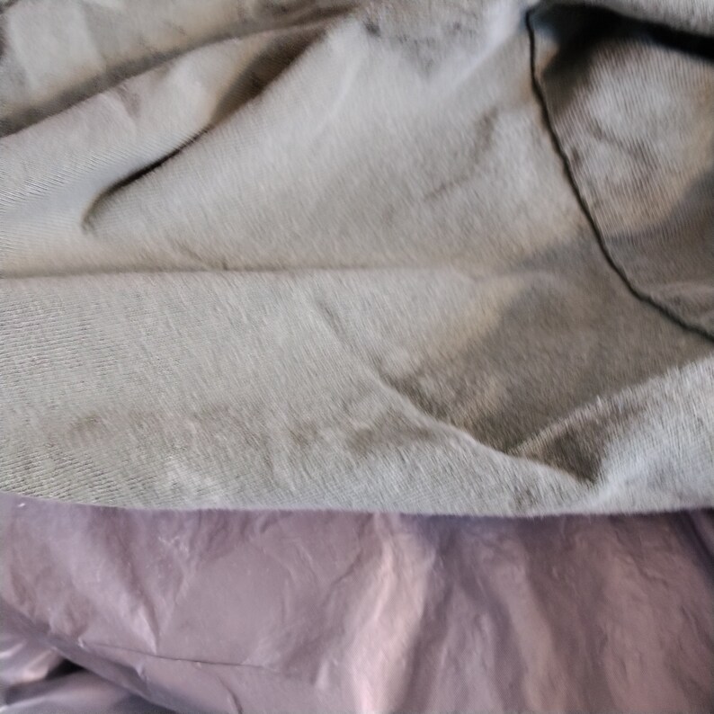 Puede incluir: Primer plano de una tela gris claro con una superficie texturizada. La tela est&aacute; doblada sobre s&iacute; misma, creando un efecto de drapeado suave.