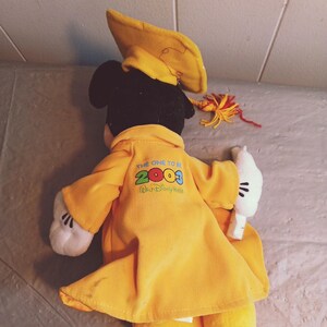 Puede incluir: Un mu&ntilde;eco de peluche de Mickey Mouse con birrete y toga amarillos. La toga tiene el texto "THE ONE TO BE 2003 Walt Disney World" impreso en la espalda. El mu&ntilde;eco tiene guantes blancos y zapatos amarillos.