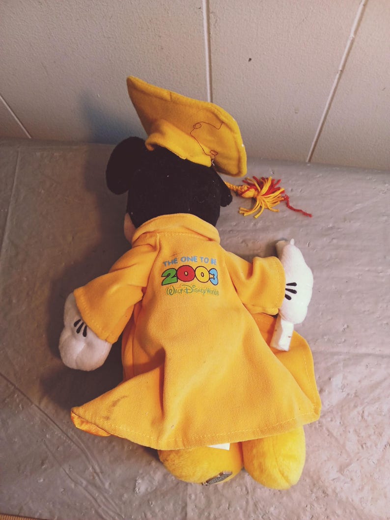 Puede incluir: Un mu&ntilde;eco de peluche de Mickey Mouse con birrete y toga amarillos. La toga tiene el texto "THE ONE TO BE 2003 Walt Disney World" impreso en la espalda. El mu&ntilde;eco tiene guantes blancos y zapatos amarillos.