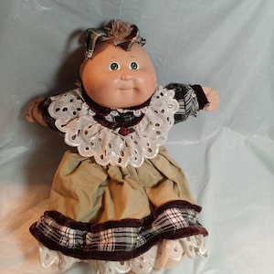 Könnte beinhalten: Eine Puppe mit rundem, gebräuntem Gesicht, grünen Augen und braunen Wollhaaren. Sie trägt ein dunkelgrün-braunes kariertes Kleid mit weißem Lochstickerei-Kragen, einem beigen Rock und einem dunkelbraunen Samtbesatz mit einem kleinen roten Akzent.