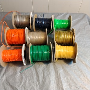 Puede incluir: Carretes de cuerda de colores en varios colores, incluyendo naranja, verde, amarillo, dorado y negro. La cuerda está enrollada alrededor de carretes de cartón.