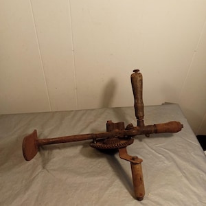 Puede incluir: Un taladro manual antiguo y oxidado, con mangos de madera y un mecanismo de engranajes. La herramienta muestra signos de edad y desgaste, lo que sugiere que es una antigüedad. Está diseñado para el funcionamiento manual, probablemente para tareas como taladrar agujeros en madera u otros materiales.