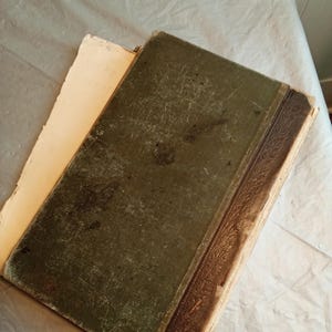 May include: An antique book with a worn, dark green cover and a brown spine. The pages are yellowed with age, and the cover shows signs of wear and tear. The book is open, revealing the aged pages.