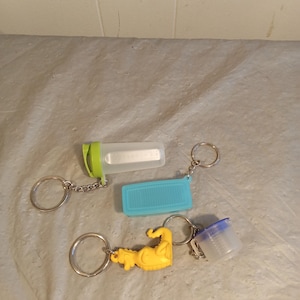 May include: A collection of keychains featuring various designs. One keychain has a clear, rectangular container with a green lid. Another has a blue, rectangular container. A third keychain has a yellow, muscular arm. The last keychain has a small, clear container with blue lid.