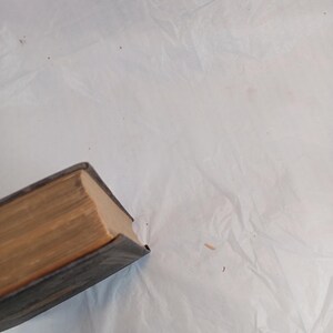May include: A partially visible, aged book with a dark blue cover and aged, tan-colored pages. The book's edges show wear, suggesting it is an antique or vintage item. The book is positioned at an angle against a white, textured background.